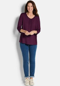 Violette Bluse mit langen Ärmeln, einem runden Ausschnitt und Faltendetails an der Vorderseite, kombiniert mit blauen Skinny Jeans und beigen Sneakern.