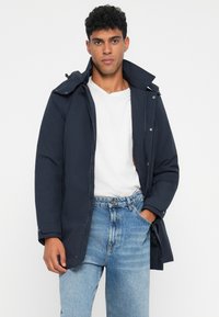 Giubbotto leggero, blu navy, con zip frontale, cappuccio e bottoni automatici. Presenta una vestibilità comoda e una texture sottile. Indossato sopra una t-shirt bianca.
