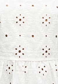 Tissu blanc avec motifs floraux brodés et petits trous découpés formant des marguerites et des feuilles décoratives.