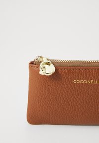 Borsetta in pelle marrone con texture, dotata di una chiusura lampo e accessori dorati. Presenta un logo impresso sul davanti. Forma compatta e rettangolare.