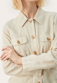Veste en lin beige clair avec col, quatre poches et boutons en bois ; texture douce avec une coupe décontractée.