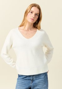 Witte, zachte gebreide trui met V-hals, lange mouwen, comfortabele pasvorm en ribgebreide onderrand. Combinatie met blauwe denim jeans voor contrast.