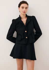 Zwarte tweed jas met franjes aan de randen, twee voorzakken en gouden knopen, gecombineerd met een bijpassende zwarte flared rok.