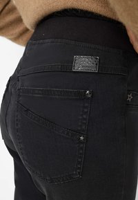 Schwarze Denim-Jeans mit glatter Textur, ausgestattet mit einem schwarzen Bund, sternenverzierten Taschenakzenten und einem Etikett an der hinteren Tasche.