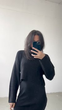 Czarny prążkowany, krótki sweter w połączeniu z czarną sukienką, z długimi rękawami i gładką teksturą. Otoczenie jest minimalistyczne.