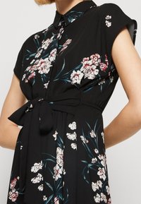 Schwarzes, kurzärmeliges Kleid mit weißem und rotem Blumenmuster, ausgestattet mit einem geknöpften Kragen, einem Taillengürtel und einem plissierten Rock.