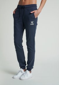 Tmavě modré tepláky z měkké látky s elastickým pasem, bočními kapsami a manžetami na kotnících; na levém stehně je bílý logo.