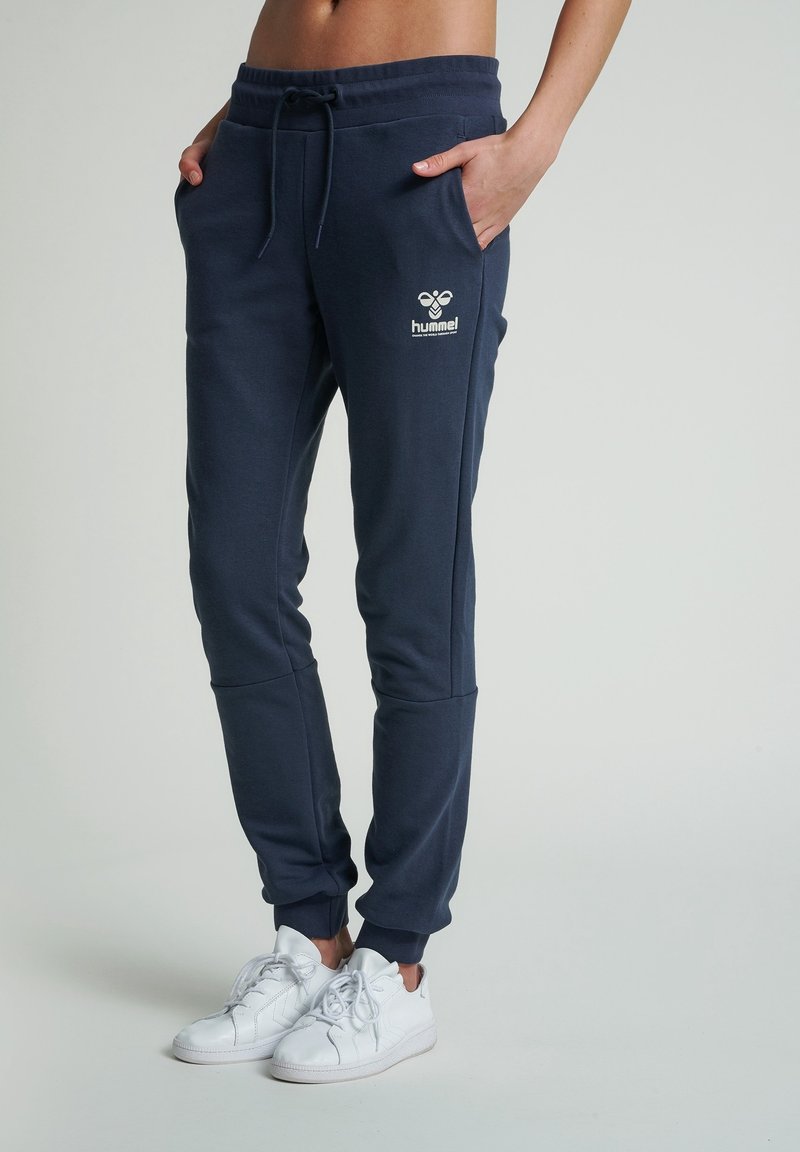 Tmavě modré tepláky z měkké látky s elastickým pasem, bočními kapsami a manžetami na kotnících; na levém stehně je bílý logo.