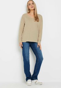 Hellbeige Langarm-Oberteil mit V-Ausschnitt, kombiniert mit blauen geraden Jeans und weißen Sneakers mit beige Akzenten. Einfaches, lässiges Outfit-Design.