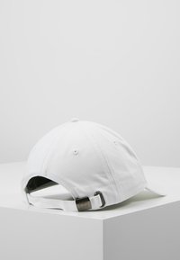 Hvid bomulds baseball cap med en buet skygge, justerbar rem bagpå og metalliske detaljer. Glat tekstur og enkelt design.