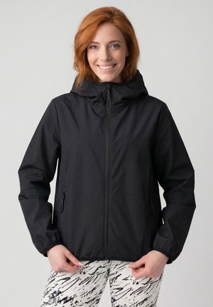 Lächelnde rothaarige Frau, die eine schwarze Kapuzenjacke mit Reißverschluss und weiße Hosen mit schwarzen abstrakten Mustern trägt, steht vor einem hellgrauen Hintergrund.