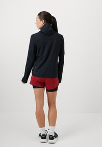 Sort hættejakke til sport med lange ærmer, tekstureret stof og tætsiddende pasform, parret med røde shorts. Minimalistisk design uden logoer.
