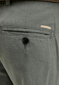 Τσέπη από πράσινο ύφασμα παντελονιού με μαύρο κουμπί και μικρή μπεζ ετικέτα «JACK & JONES» κοντά στην πάνω άκρη.