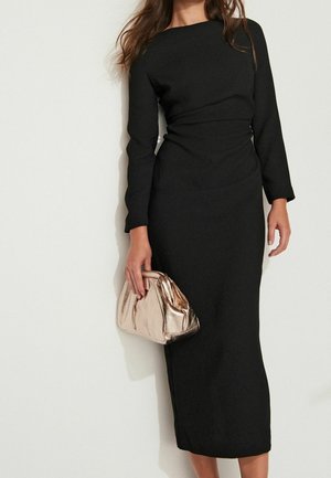 Kobieta w czarnej sukience midi z długim rękawem, trzymająca metaliczną złotą kopertówkę na jednolitym jasnym tle.