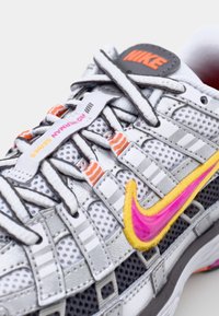 Zapatilla Nike con una parte superior de malla gris, acentos multicolores, un swoosh rosa vibrante y cordones con patrón. Suela de goma gruesa con tracción.