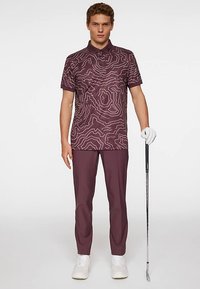 Golfpiké i mörklila med ett vitt konturmönster, kombinerad med matchande byxor, vita golfsko och en golfklubba.