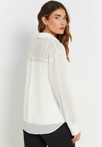Blusa bianca e trasparente con colletto, maniche lunghe e polsini con bottoni. Presenta una parte inferiore opaca in contrasto e semplici dettagli di cucitura.