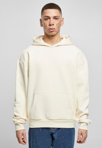 Sudadera crema con un bolsillo canguro en la parte delantera, capucha con cordones y puños acanalados, llevada sobre unos vaqueros azul denim, con un ajuste holgado.