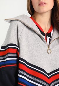 Mujer con una sudadera gris con cremallera dorada y franjas de colores rojo, azul, negro y blanco en las mangas y el pecho, labios ligeramente entreabiertos.