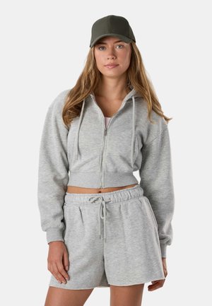 Young woman wearing a zipped grey cropped hoodie, matching drawstring shorts, and an olive green baseball cap, standing against a white background.