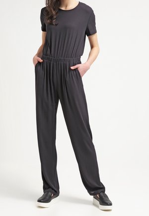 Schwarzer Jumpsuit mit lockerer Passform, kurzen Ärmeln, elastischem Taillenbund und seitlichen Taschen, kombiniert mit schwarzen Slippern und flacher Sohle.