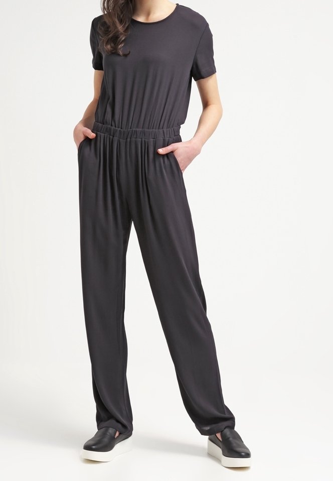 Musta haalari rento leikkauksella, lyhyet hihat, joustava vyötärö ja sivutaskut, yhdistettynä mustiin liukulaitoskenkiin, joissa on litteät pohjat.