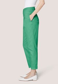 Grüne, konisch geschnittene Hose mit vorderen Falten, Seitentaschen und glatter Textur, kombiniert mit weißen Ballerinas. Das Design ist lässig geschnitten und knöchellang.