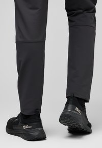 Schwarze Sportschuhe mit einer strukturierten Sohle und gepolstertem Absatz, kombiniert mit eng geschnittenen, dunkelgrauen Hosen aus glattem, dehnbarem Material.