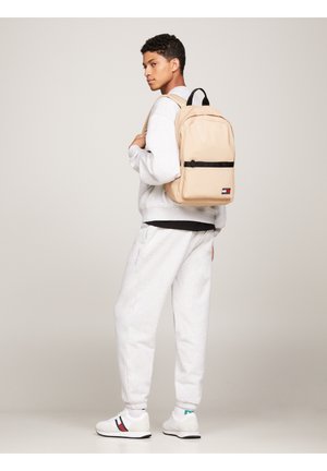 Zaino beige con accenti neri, forma rettangolare e più scomparti. Indossato con una felpa grigia e pantaloni della tuta abbinati.