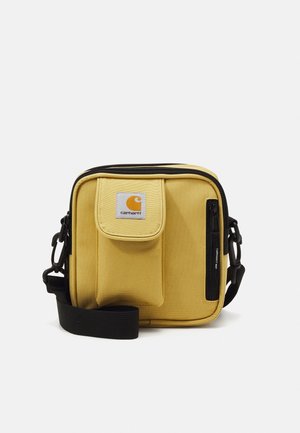 Bolso bandolera Carhartt amarillo con bolsillo delantero de solapa, bolsillo lateral con cremallera, correa de hombro negra ajustable y parche de logo visible.