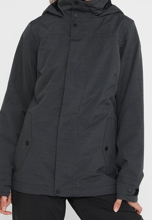 Persoon draagt een donkergrijze capuchonjas met drukknopen aan de voorkant en ritszakken aan de zijkant, gekleed in een zwarte broek.