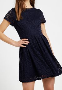Námořnická modrá krajková šaty s krátkými rukávy, fitted živůtek a rozšířená sukně. Obsahují květinový vzor a detaily s vlnkovým lemem.