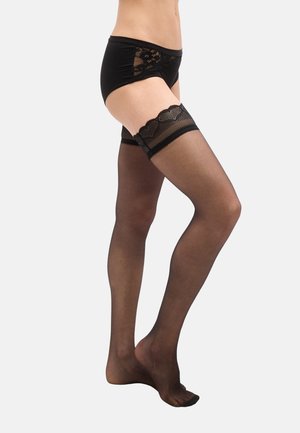 Donna che indossa calze autoreggenti nere trasparenti con pizzi sulla parte superiore e biancheria intima coordinata rifinita in pizzo nero, in piedi contro uno sfondo bianco.
