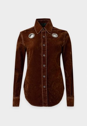 Mujer con cabello largo y rizado que lleva una camisa y pantalones de pana marrón decorados con ojales metálicos redondos, top blanco corto y botas negras.