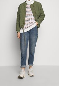 Grüne gesteppte Bomberjacke über einem mehrfarbigen gemusterten Pullover, kombiniert mit blauen Jeans mit umgeschlagenem Saum und weißen Sneakers.