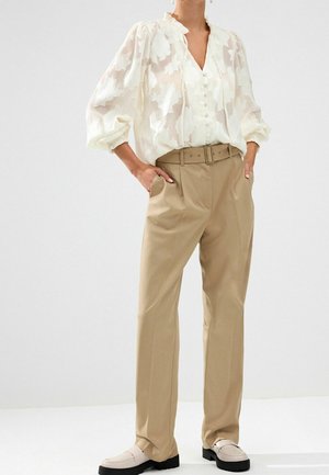 Femme portant un chemisier blanc transparent à motifs floraux avec des manches bouffantes, un pantalon beige à taille haute ceinturé, et des mocassins crème, les mains dans les poches.