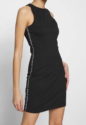 Kobieta nosząca czarną sukienkę bez rękawów z białym napisem "Calvin Klein Jeans" biegnącym pionowo wzdłuż bocznego szwu.