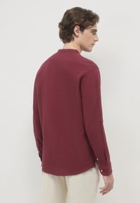 Uomo in una camicia bordeaux a maniche lunghe con polsini abbottonati, girato leggermente di lato, indossando pantaloni beige chiaro.