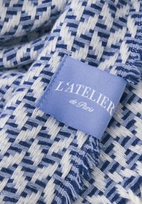 Tkany materiał z niebiesko-białym wzorem przeplatanym oraz widoczną metką z napisem "L'Atelier de Paris" w białej czcionce na jasnoniebieskim tle.