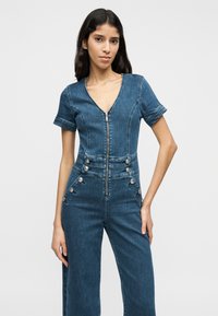 Ολόσωμη φόρμα από τζιν με V-λαιμόκοψη, κοντά μανίκια, φερμουάρ μπροστά και διακοσμητικά κουμπιά στα πλαϊνά. Σκούρο μπλε χρώμα και δομημένη εφαρμογή.