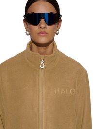 Vrouw met grote zwarte wraparound-zonnebril, zilveren creolen en een beige fleecejas met rits met "HALO" in reliëf op de borst.