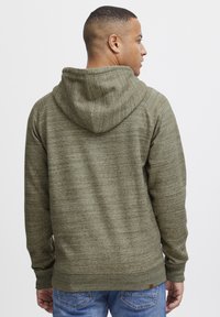Sudadera verde oliva con capucha y puños texturizados y con acanalado. Tiene un corte relajado y se combina con jeans de mezclilla azul.