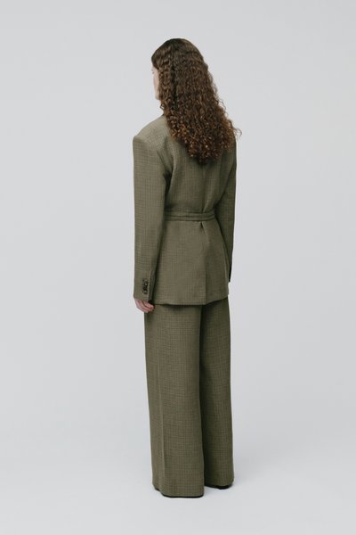 Femme aux longs cheveux bouclés, debout de dos, portant une veste de costume à carreaux vert olive avec un pantalon large assorti.