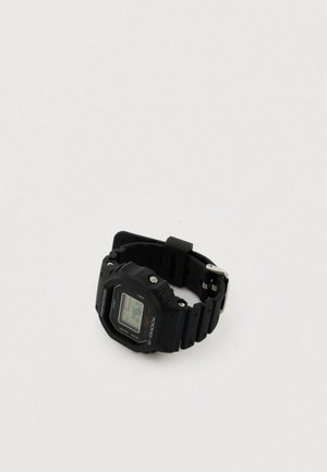 Fekete digitális óra gumipánttal, négyzet alakú számlappal, LCD kijelzővel és fém alkatrészekkel. A címkén "G-SHOCK" felirat és különböző műszaki adatok találhatók.