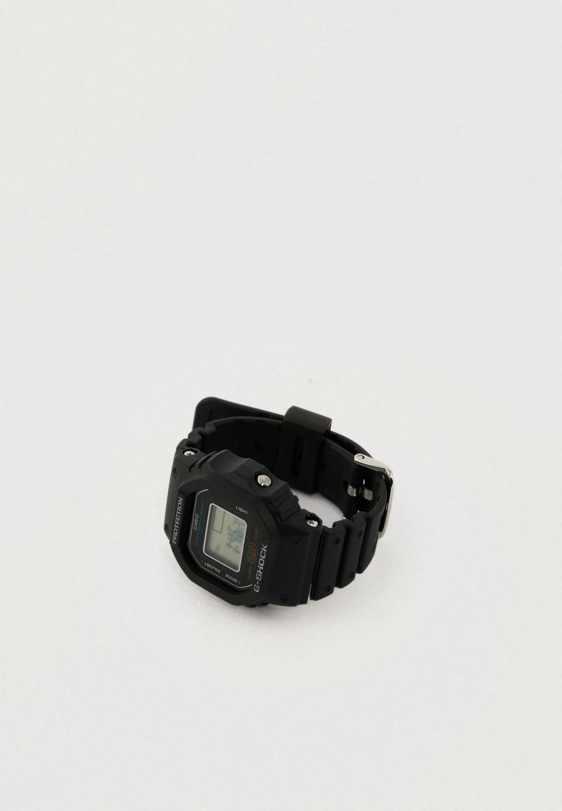 Fekete digitális óra gumipánttal, négyzet alakú számlappal, LCD kijelzővel és fém alkatrészekkel. A címkén "G-SHOCK" felirat és különböző műszaki adatok találhatók.