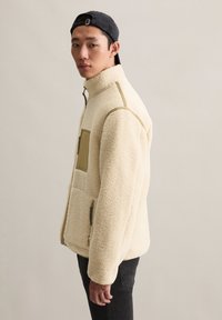 Fleece jack in off-white met een gestructureerd, zacht oppervlak, voorzien van een voorzak met een rits en groene accenten.