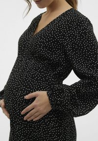 Črna obleka s belimi pikami, ki ima dolge rukave, V-izrez in elastičen pas, prilagojen za nosečniški silhuet.