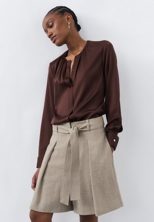 Kvinde iført en mørkebrun bluse med lange ærmer og højtaljede beige shorts med bælte, stående foran en hvid baggrund.