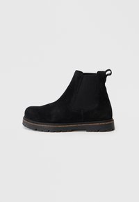 Stivale Chelsea anklet in suede nero con pannello elastico laterale, linguetta sul tallone e suola robusta con rifinitura marrone su sfondo semplice.