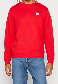 Sudadera roja de manga larga con puños acanalados, con un pequeño parche de logo azul en el pecho. Combina con pantalones vaqueros azules.